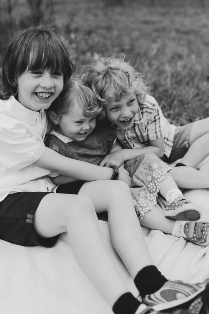 three siblings are sitting on the blanket and laughing. black and white natural lifestyle photo shoot in Hampshire. Hampshire family photographer. Ewa Jones Photography