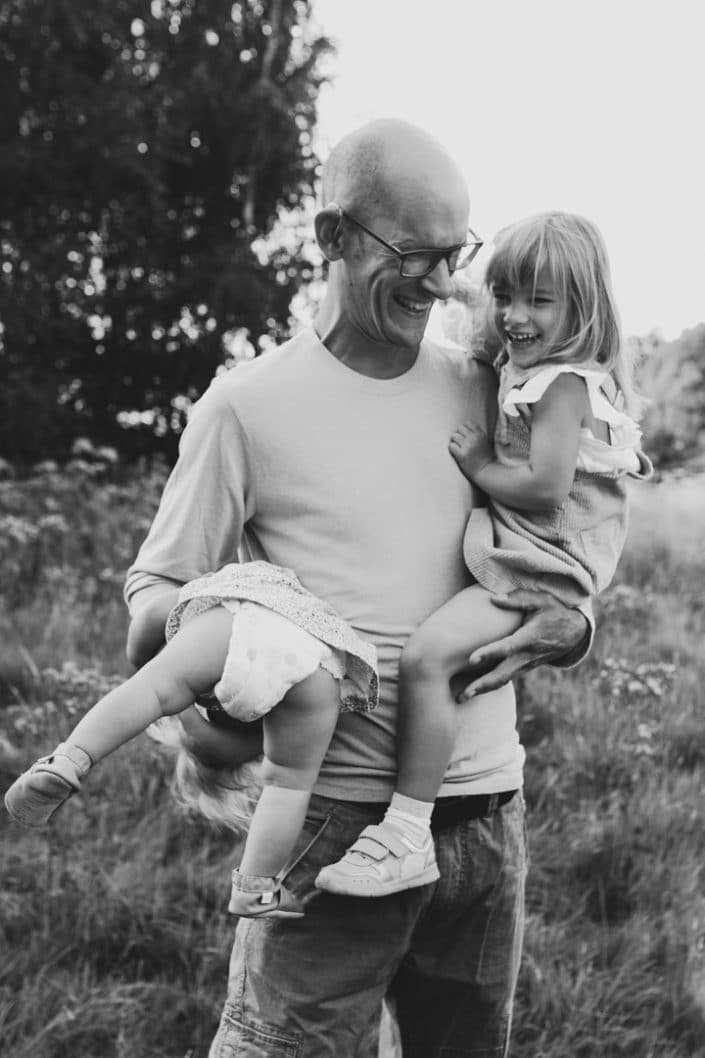 Dad is holding two daughters on his arms. One daughter is upside down and another is laughing. Family photographer in Hampshire. Ewa Jones Photography