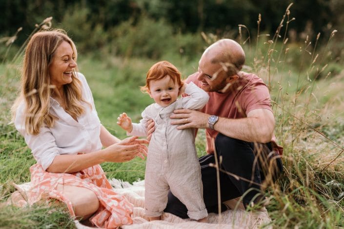 Mum and dad are sitting on the blanket and boy is standing and laughing. Family photographer in Hampshire. Mum is standing in the field and holding her newborn boy. Lovely natural family photo session in Hampshire. Basingstoke photographer. Ewa Jones Photography