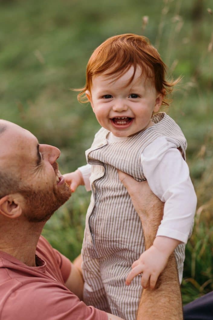 Little boy is looking at the camera and smiling and dad is looking at him. Unposed natural family photo session in Hampshire. Mum is standing in the field and holding her newborn boy. Lovely natural family photo session in Hampshire. Basingstoke photographer. Ewa Jones Photography