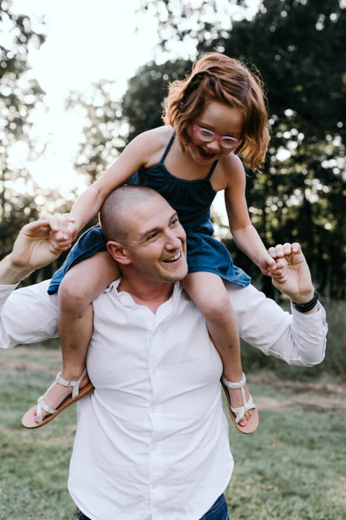 Daughter is on daddy's shoulders and laughing. Shoot in front of the sun back lit. Natural family photoshoot in Hampshire. Ewa Jones Photography