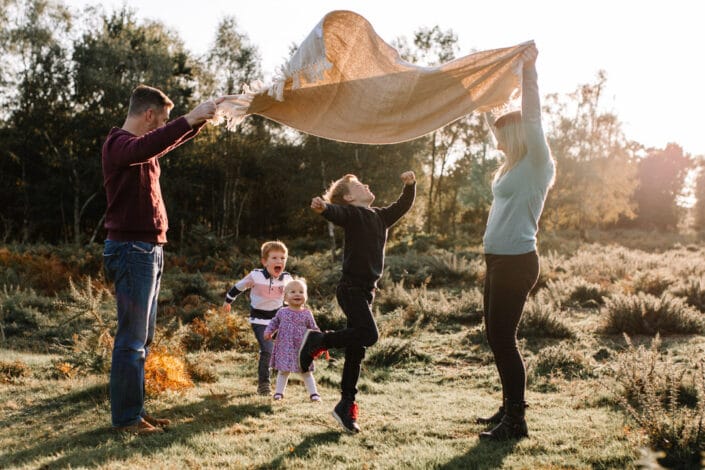 Mum and dad are holding a blanket and lifting it in the air. Three children are running through the blanket. Autumn family photo session in Hampshire. Ewa Jones Photography