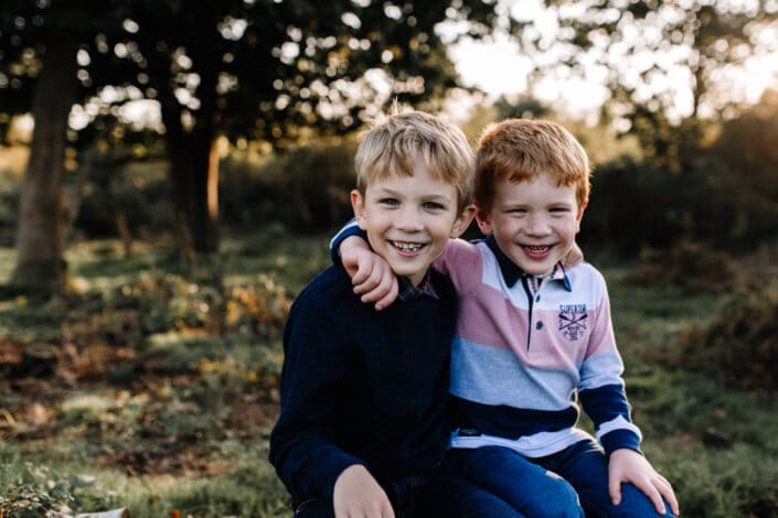 Two brothers are sitting on the log and looking at the camera. They are both giggling. One boy is wearing blue jumper and one is wearing stripy top. Autumnal family photo session in Hampshire. Family photographer in Hampshire. Ewa Jones Photography