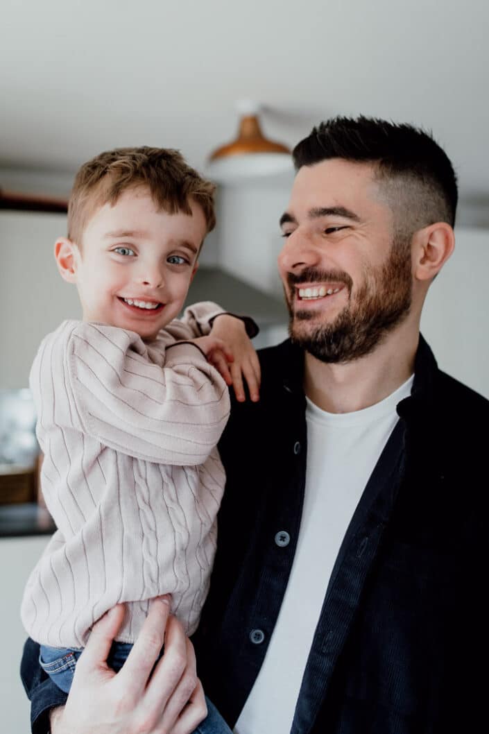 Dad is holding his son and looking at him lovingly. Son is smiling and wearing cream jumper. Izzo family photo session. Family lifestyle photography in Hampshire. Ewa Jones Photography