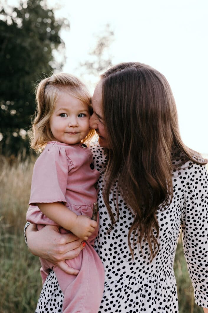 Mum is standing and holding her daughter. she is smiling to her. Mum is wearing lovely white dress with black dots and girl is wearing pink jump suit. Family lifestyle photography in Basingstoke, Hampshire. Ewa Jones Photography