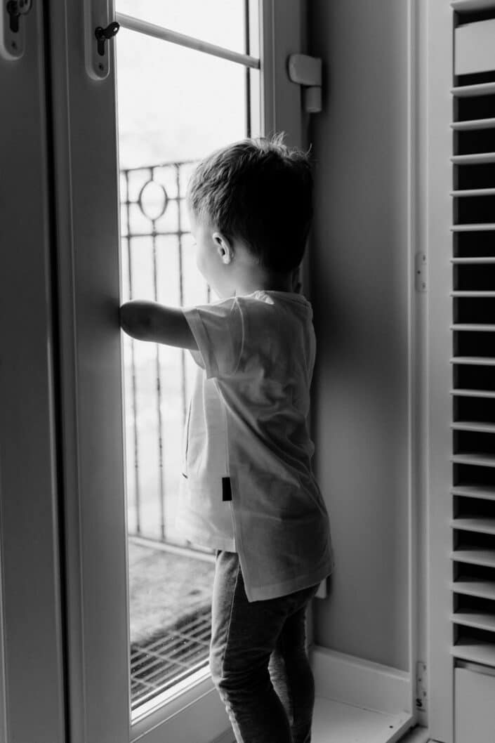 Black and white photograph of little boy looking through the window. Family in home lifestyle photography in Basingstoke. Ewa Jones Photography