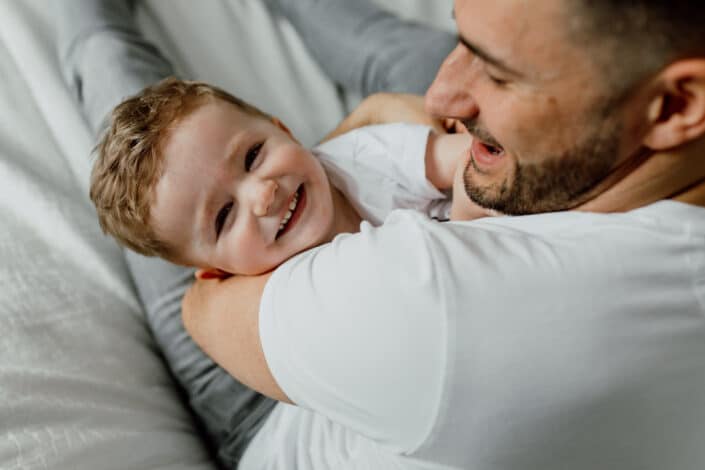 Dad is cuddling tight his little son and son is looking and laughing into the camera. Family lifestyle photography in Hampshire. Ewa Jones Photography