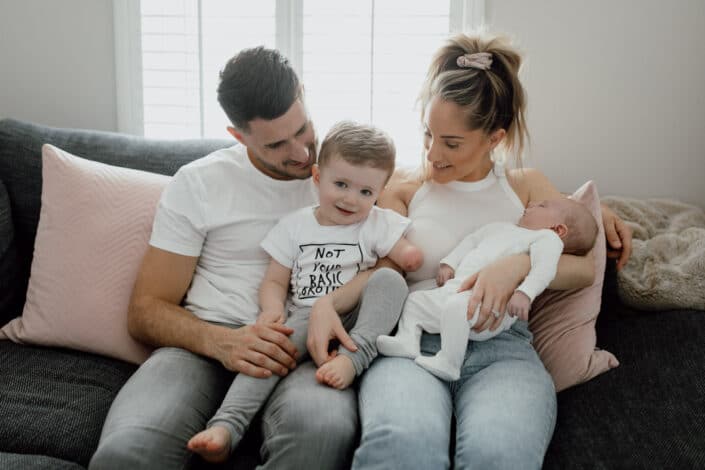 Mum, dad and two sons are sitting on the sofa and smiling. All are wearing white t-shirts and jeans. Lovely family in home lifestyle photography in Basingstoke. Ewa Jones Photography