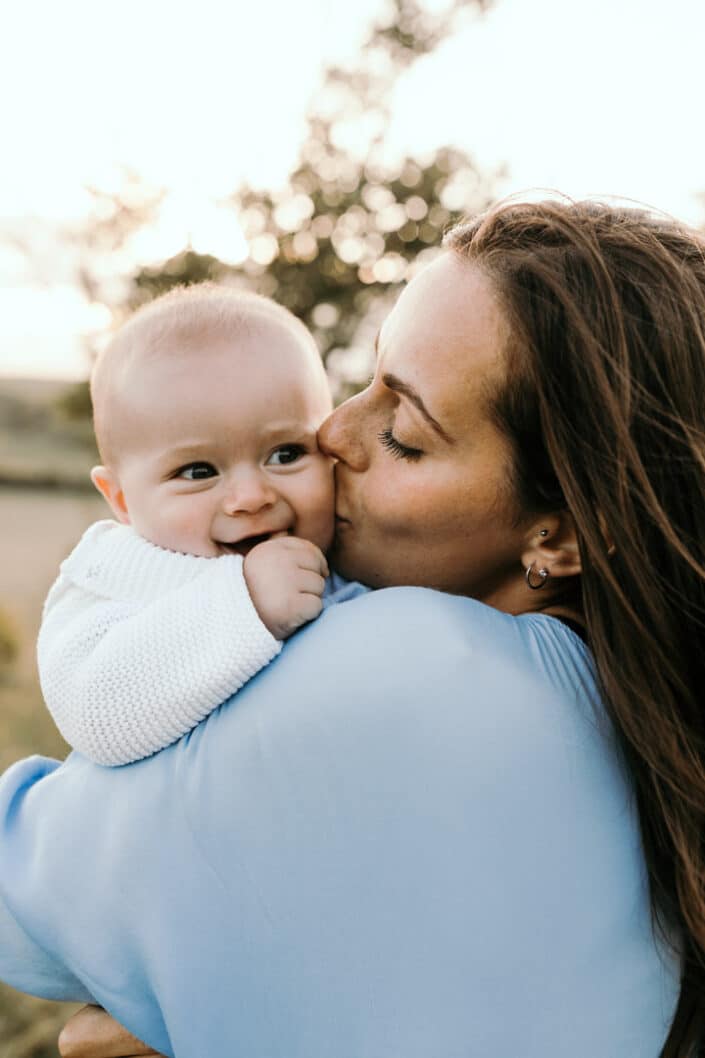 Mum is kissing her little boy on the cheek. She is wearing lovely blue dress and boy is wearing a white cardigan. Family photography in Salisbury. Ewa Jones Photography