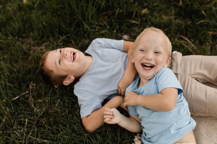 Two brothers are laying on the grass and laughing. Both boys are wearing blue t-shirts. Lovely family photography in Hook, Hampshire. Ewa Jones Photography