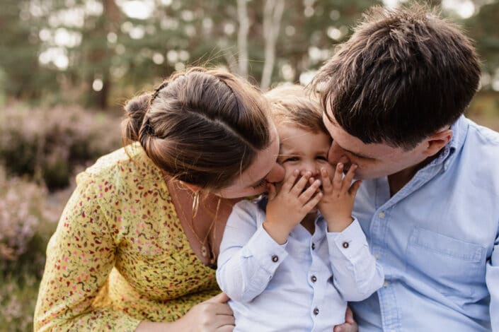 Mum and dad are kissing their little son on his cheeks. Boy is covering his mouth with his hands and is giggling. Family lifestyle photo session in Hampshire. Ewa Jones Photography