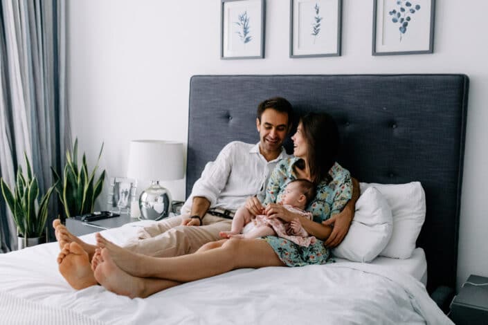 Mum, dad and their little baby girl are sitting on the bed. Mum and dad are looking lovingly at each other. Lovely natural family photography in Hammersmith. Ewa Jones Photography