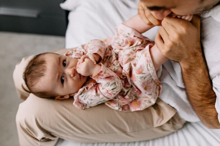 Dad has his little girl on his knees. She is laying down and sucking her thumb. Natural lifestyle photo session In Hammersmith, London. Ewa Jones Photography