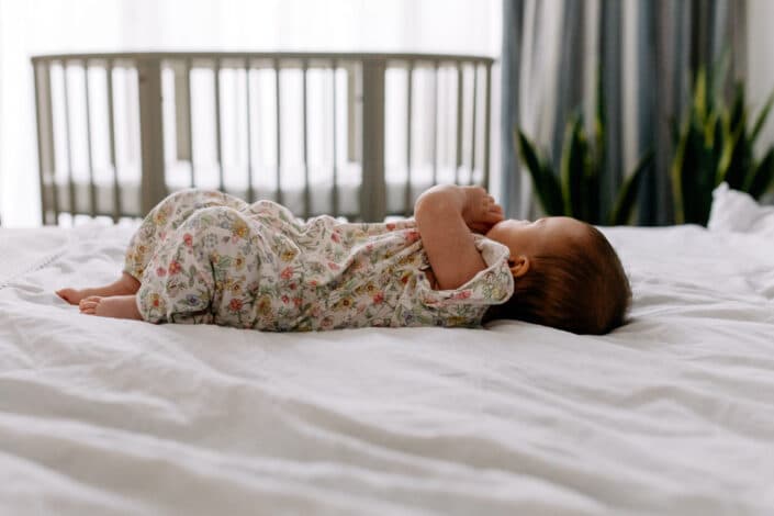 Little baby girl is laying on the bed and looking away. Baby cot in the background. Baby girl is wearing flower body. Family photography in London. Ewa Jones Photography