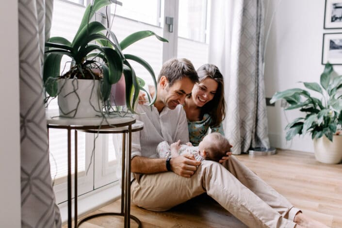 Mum and dad are sitting on the floor and looking at their baby girl. There are plants in front of them and lovely light coming from the outside. Natural photography. Ewa Jones Photography