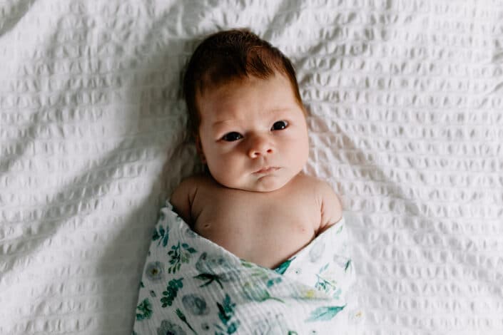 Gorgeous newborn baby girl is laying on the bed and looking up at the camera. She is wrapped in a baby blanket. lifestyle photography in Farnham. Ewa Jones Photography