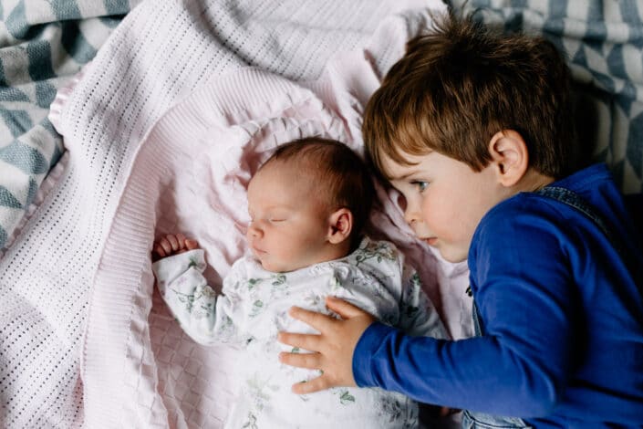 Little baby girl is laying on the blankets and sleeping. Her older brother is looking at her and holding his hand on her chest. Lovely lifestyle newborn baby photography in Hampshire. Ewa Jones Photography