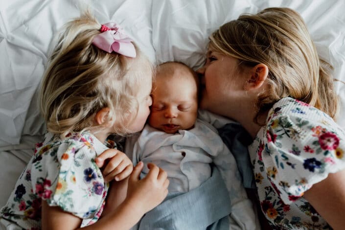 Two sisters are laying on the bed and kissing their baby brother. Girls are wearing lovely flower dresses and baby boy is wearing baby grow. Natural unposed newborn baby photography in Basingstoke, Hampshire. Ewa Jones Photography