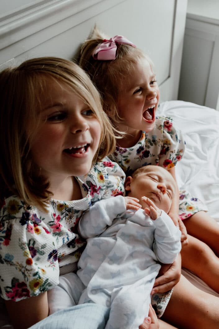 Two older sisters are holding their newborn baby boy in their arms. Lovely natural moment of family happiness. Siblings love. Newborn photographer in Basingstoke, Hampshire. Ewa Jones Photography