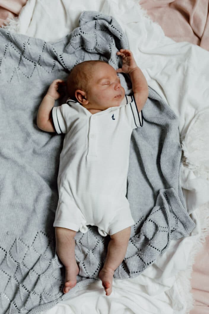 Newborn baby boy is laying on throws and sleeping. He has his hands up and he is wearing white baby grow. Newborn photography poses. Newborn photographer in Basingstoke, Hampshire. Ewa Jones Photography