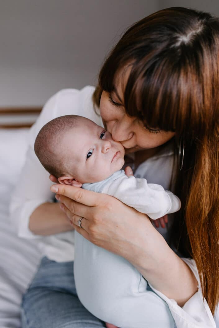 Mum is kissing her newborn baby boy. He is wearing lovely blue baby grow. Newborn baby photography in Reading, Berkshire. Ewa Jones Photography
