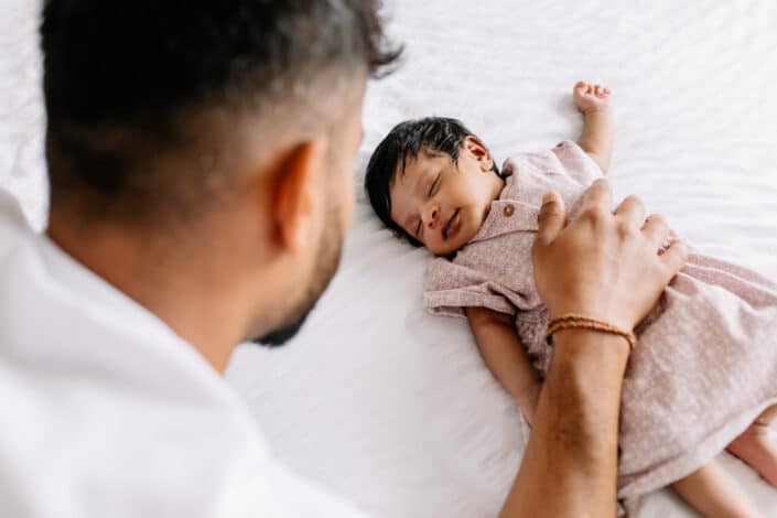 Newborn baby girl is sleeping on bed and smiling. Dad has one hand on the newborn baby girl chest. Natural lifestyle photography in London. Ewa Jones Photography