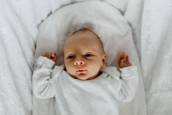 Baby girl is laying in the moses basket and looking up at the camera. She is wearing white baby grow. natural lifestyle newborn baby photographer in Basingstoke, Hampshire. Ewa Jones Photography