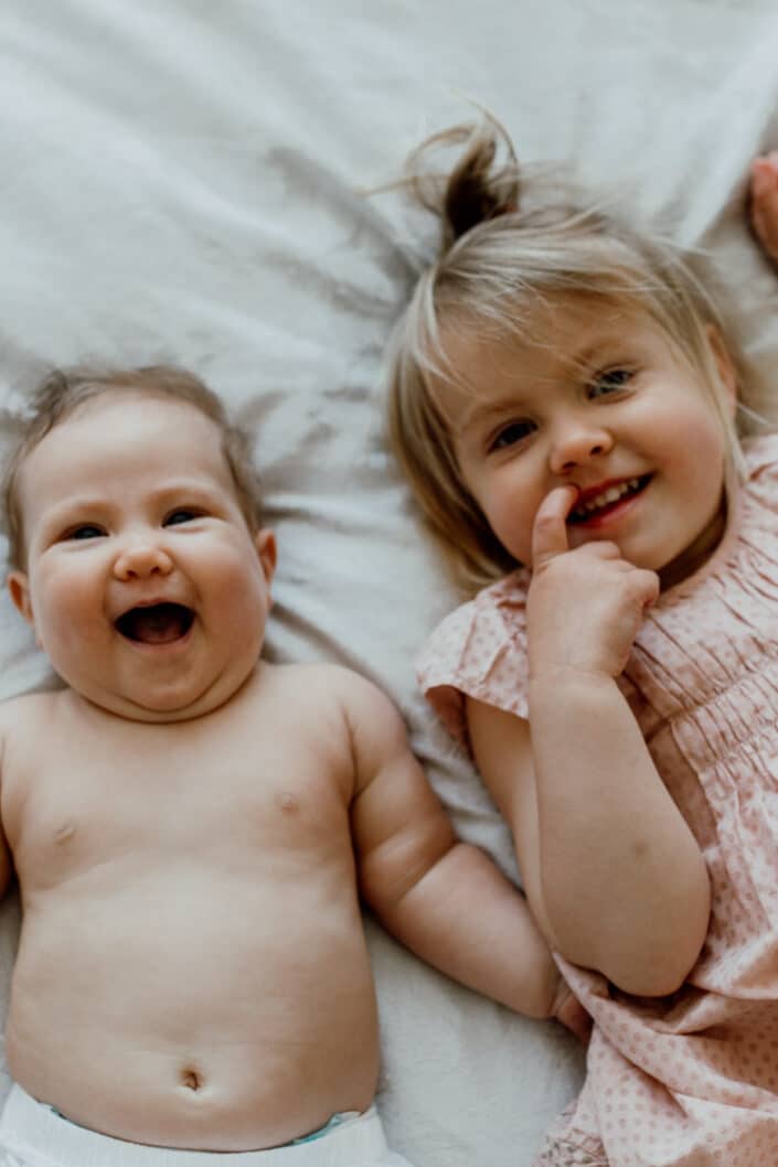 Baby girl and her older sister are laying on bed and smiling. Lovely natural lifestyle newborn photography in Hampshire. Mum and dad are cuddling close to each other and mum is holding her newborn baby girl. Lifestyle newborn photography in Hampshire. Ewa Jones Photography