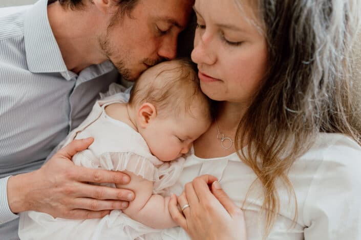 Mum and dad are cuddling close to each other and mum is holding her newborn baby girl. Lifestyle newborn photography in Hampshire. Ewa Jones Photography