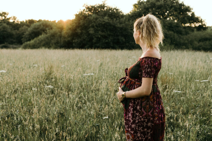 Pregnant mum is standing in the long grass and looking into the sun. Maternity photo shoot during sunset. Ewa Jones Photography