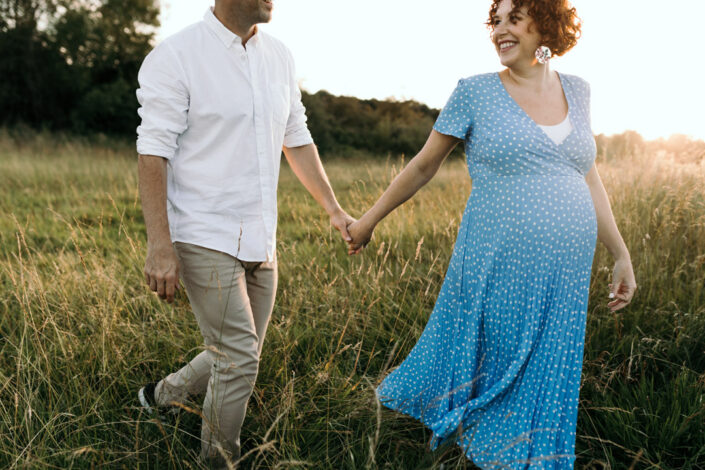 Expecting parents are holding hands. Wife is first and looking back at her husband and smiling to him. Maternity photo session in Hampshire. Ewa Jones Photography