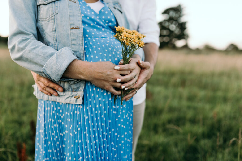 Expecting parents are holding bump and mum is holding yellow flowers. Lovely sunset maternity photo shoot in Hampshire. Ewa Jones Photography