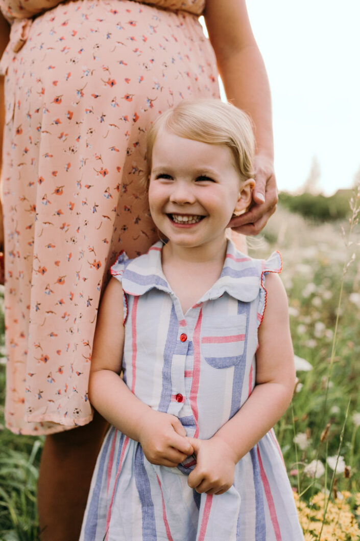 Little girl is standing and smiling. She is wearing a stripy dress. Pregnant mum is standing behind her and touching her hair. Maternity and family photography in Basingstoke. Ewa Jones Photography