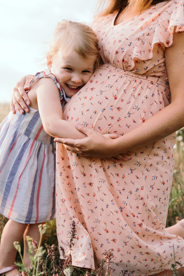 Little girl is cuddling bump. Mum is wearing lovely salmon dress. Maternity photo shoot in Basingstoke. Ewa Jones Photography