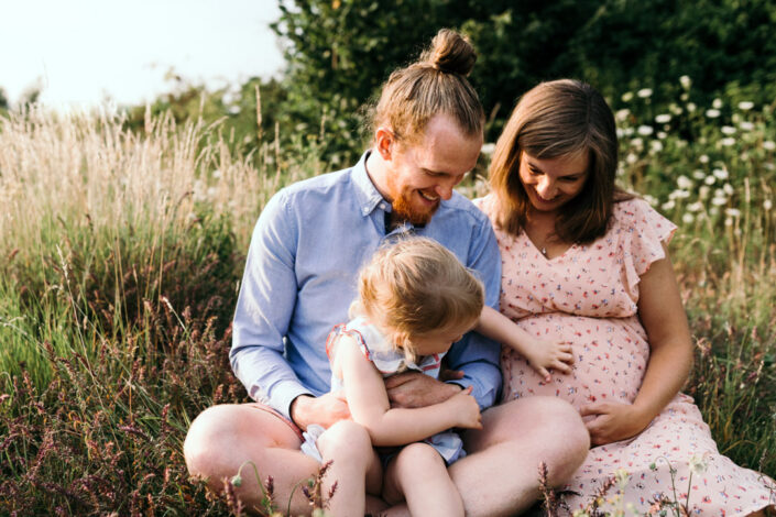 Parents and little girl are sitting on the grass. Little girl is touching her mum bump. Maternity and family photographer in Basingstoke. Ewa Jones Photography