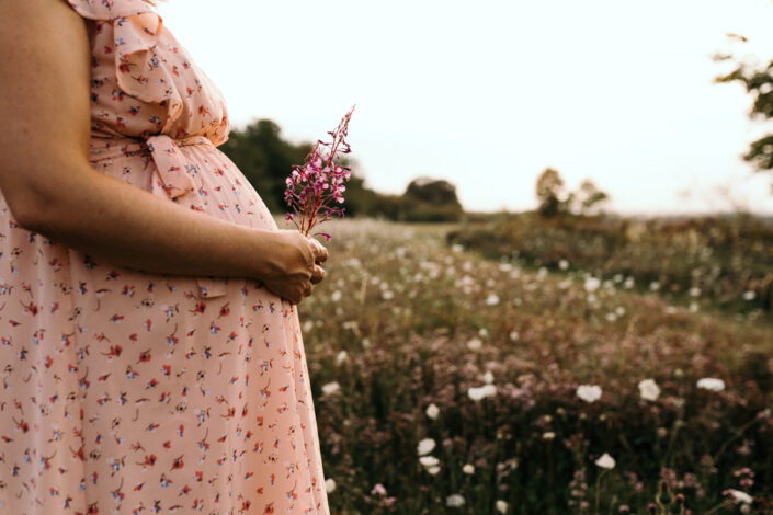 Pregnant mum is wearing a lovely dress and holding pink flowers. She is standing on a lovely field. Maternity photographer in Hampshire. Ewa Jones Photography