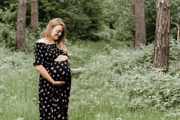 Pregnant mum is holding her bump and standing in the grass. Behind here there is lovely forest. Maternity photography in Hampshire. Ewa Jones Photography