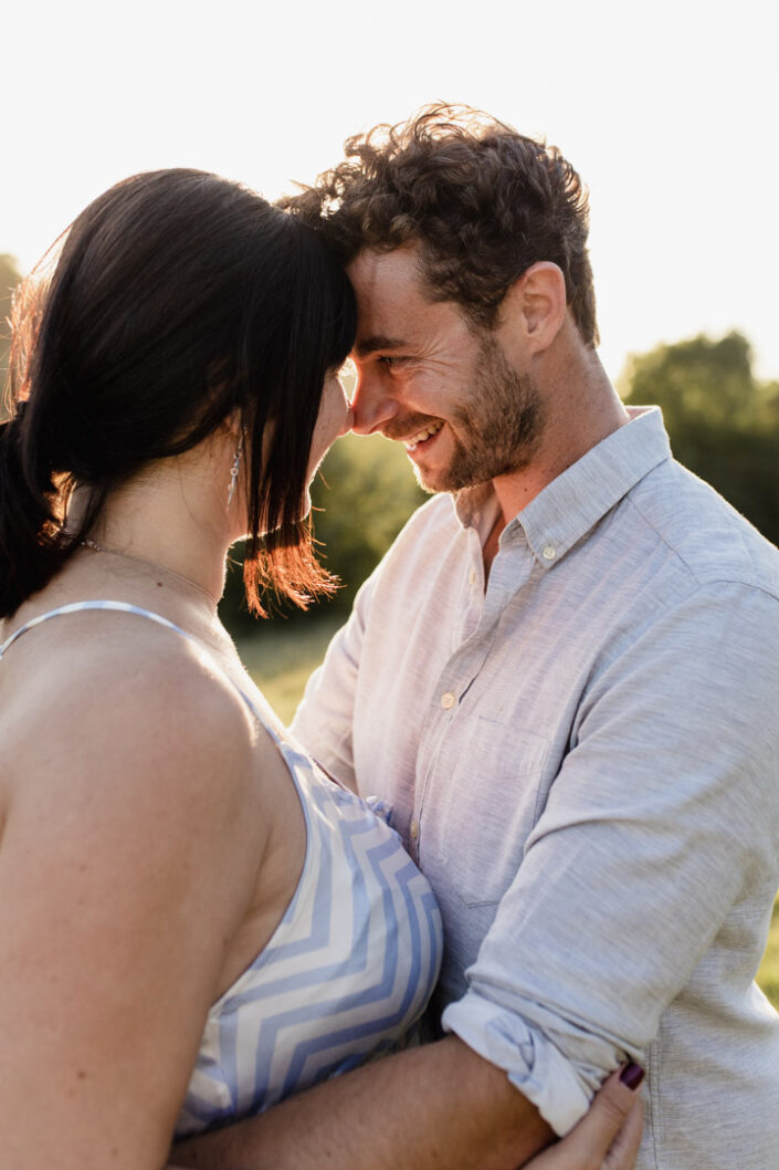 Dad is looking lovingly at his partner. Both are touching foreheads and smiling to each other. Sunset maternity photo session. Ewa Jones Photography