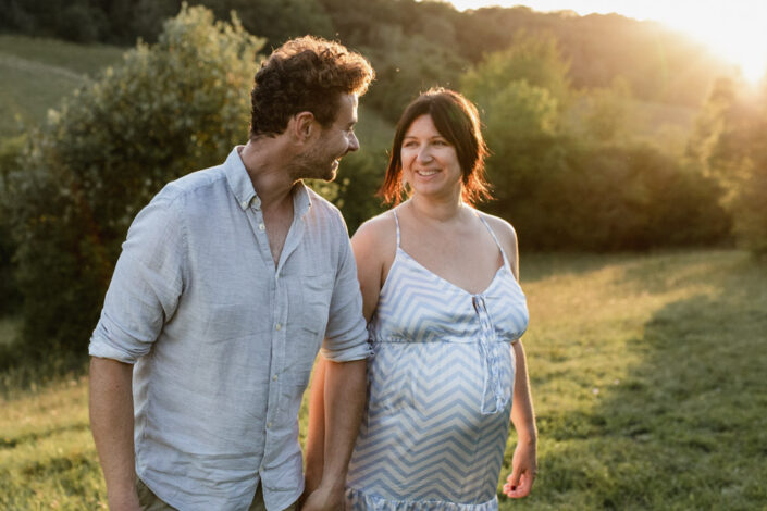 Parents to be are holding hands and looking at each other. They are walking and the lovely sunset is behind them. Natural lifestyle maternity photography in Hampshire. Ewa Jones Photography