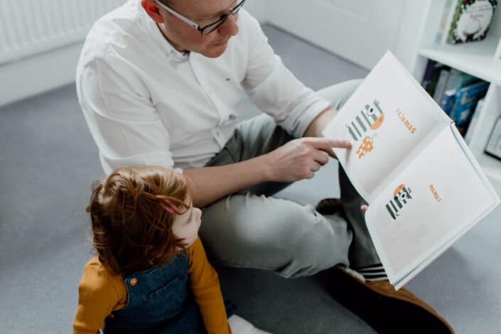 Dad is sitting on the floor and reading book to his toddler daughter. Family photographer in Basingstoke, Hampshire. Ewa Jones Photography