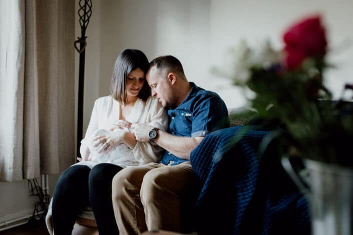 Mum and dad are sitting on the sofa and looking at their newborn baby. Newborn photographer in Basingstoke, Hampshire. Ewa Jones Photography