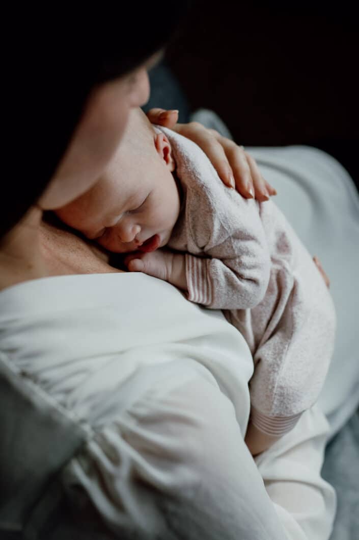 Mum is sitting on the edge of the bed and cuddling her newborn baby girl. Baby girl is sleeping and mum placed her left hand on newborn baby back. Newborn photographer in Hampshire. Ewa Jones Photography
