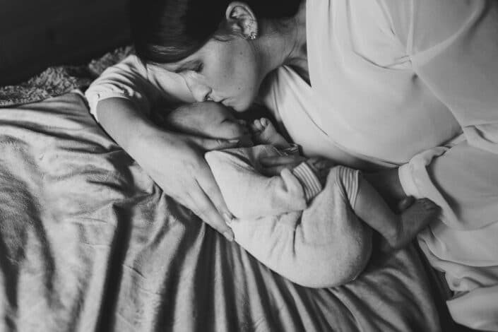 Mum is laying on the bed and she has her arm wrapped around the baby. Baby is cuddled to her mum. Mum is kissing baby on her head. Newborn baby photography in Basingstoke, Hampshire. Ewa Jones Photography