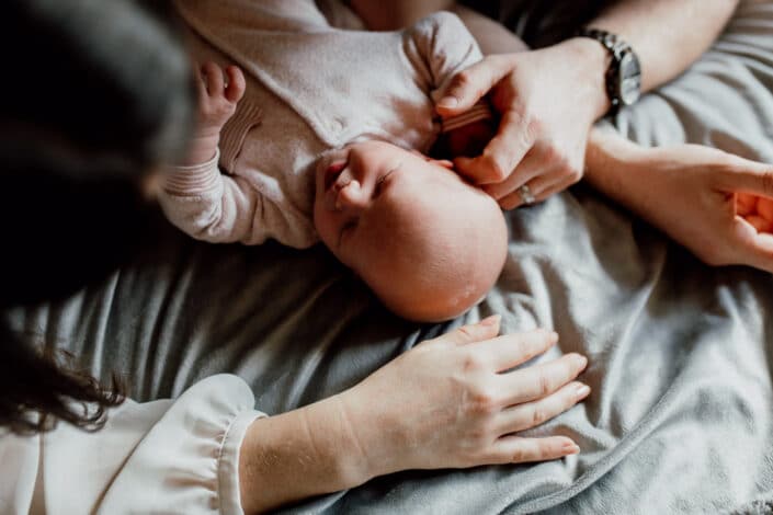 Newborn baby is laying on the bed and mum and dad have their hands close to newborn baby head. Lovely candid shoot during newborn lifestyle photo session in Basingstoke, Hampshire. Ewa Jones Photography