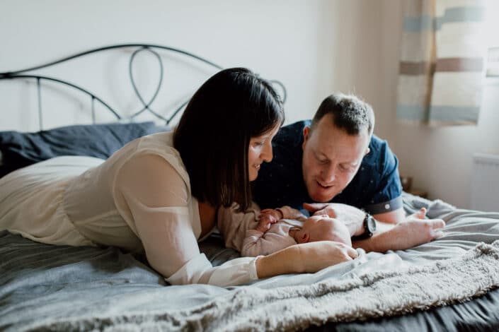 Parents of the newborn baby are laying on the bed on their tummy's. They are looking at their beautiful newborn baby girl and smiling to her. Newborn photographer in Hampshire. Ewa Jones Photography