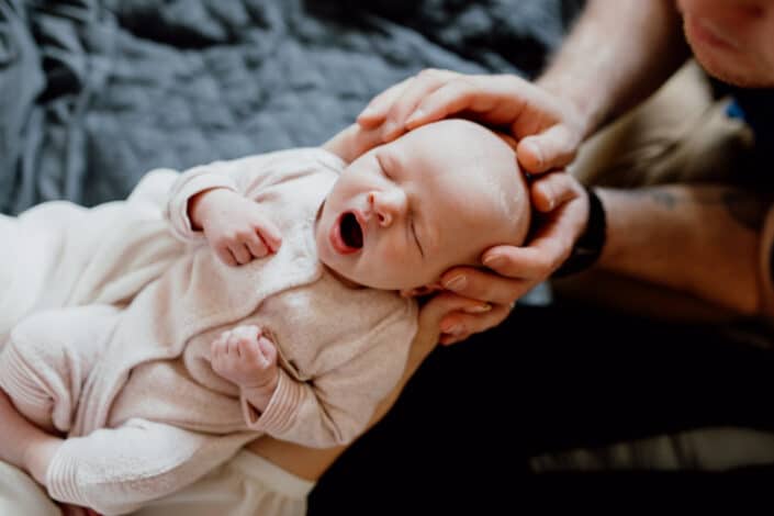 Little newborn baby girl is yawning. Dad has his hands placed on his baby girl head. Newborn baby photo shoot in Basingstoke, Hampshire. Ewa Jones Photography