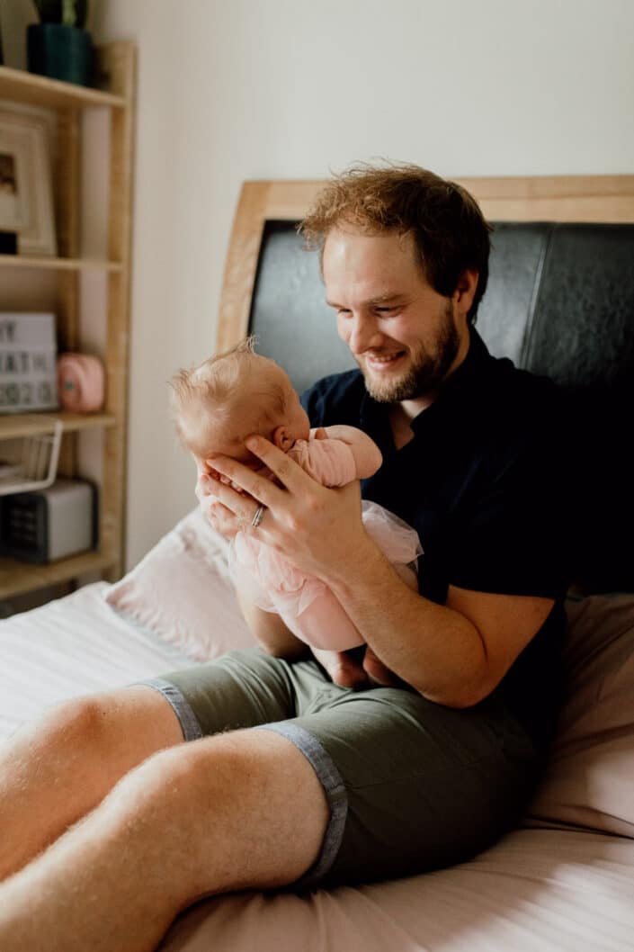 Dad is sitting on the bed and holding his little newborn baby girl. He is smiling to her and baby girl is wearing a pink baby dress Newborn photography in Hampshire. Ewa Jones Photography