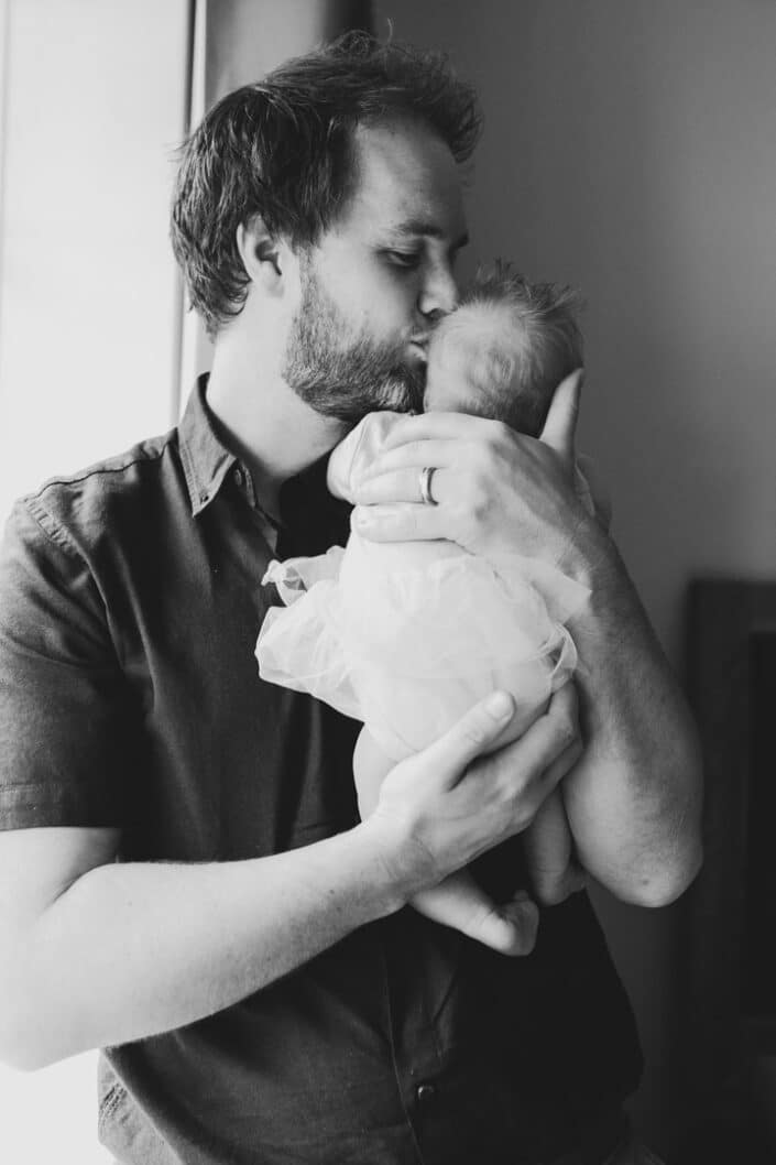 Dad is kissing his newborn baby girl on her head. He is standing next to the window and the picture is in black and white. Newborn photo session in Hampshire. Ewa Jones Photography