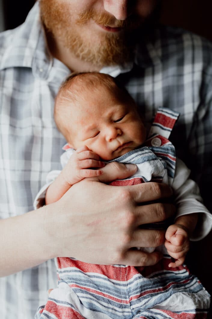 Dad is standing next to the window and holding his newborn baby. Baby boy is sleeping and enjoying dadd's cuddles. Newborn photo session in Basingstoke, Hampshire. Newborn baby feet detail shoot. Newborn baby is holding his feet together whilst laying on the bed. Newborn baby photography in Hampshire. Ewa Jones Photography