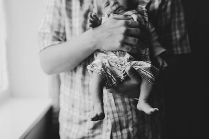 Dad is holding newborn baby. Details of the newborn baby feet, Newborn in-home family session in Hampshire. Newborn baby feet detail shoot. Newborn baby is holding his feet together whilst laying on the bed. Newborn baby photography in Hampshire. Ewa Jones Photography